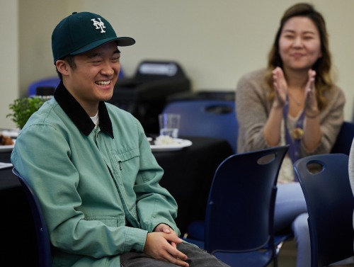 A male student wearing a Yankees baseball hat and green jacket smiles as a woman claps in the background