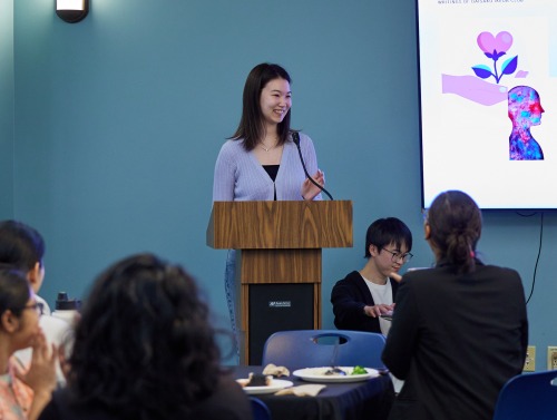 A woman smiles as she stands at the podium and presents during a seminar series