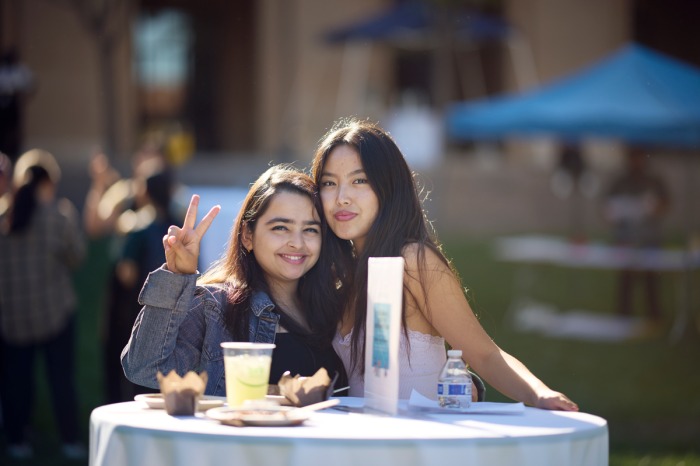 Two female students smile for a photo as they stand at an outdoor table.