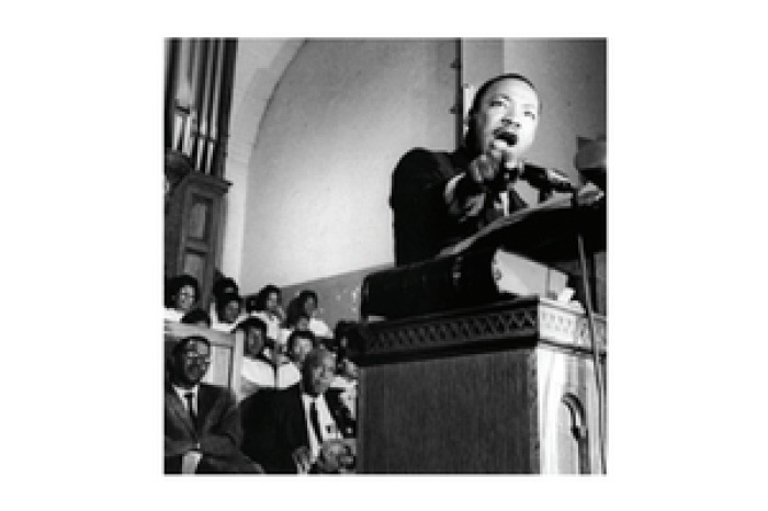 A black and white photograph depicting Martin Luther King Jr. speaking passionately into a microphone from a podium with a group of people standing behind him.