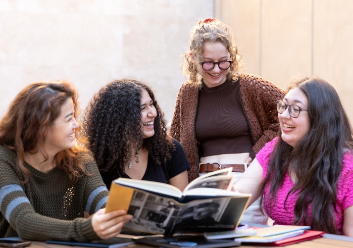 Four female Humanities students share a laugh while looking at a book together.