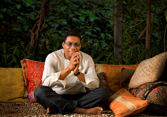 Herbie Hancock sits crosslegged surrounded by colorful throw pillows. He is clasping his hands and smiling at the camera while wearing a white striped shirt, dark jeans, and tinted glasses. Photo by Douglas Kirkland