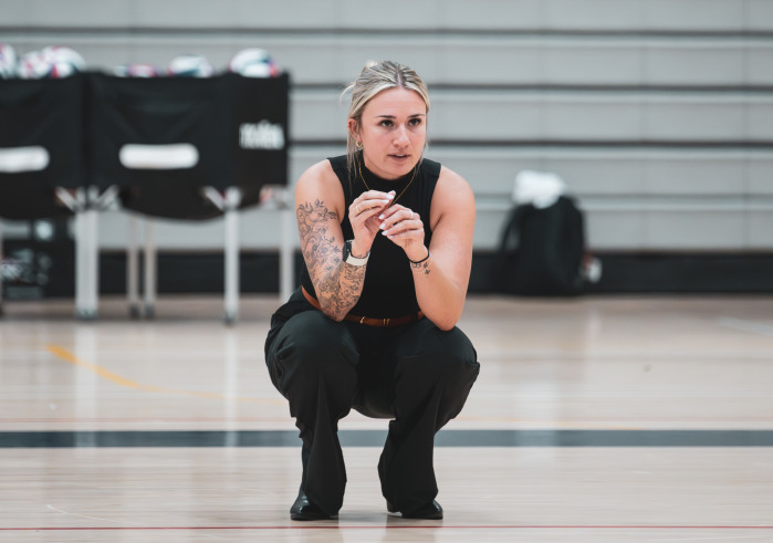 A blonde woman with her hair up, wearing a black tanktop and pants squats down on a gym floor with volleyballs in the distance.