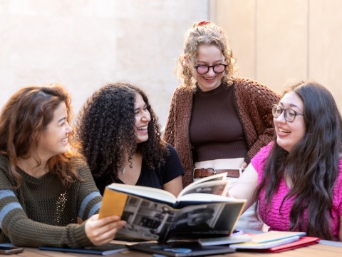 Four female Humanities students share a laugh while looking at a book together.