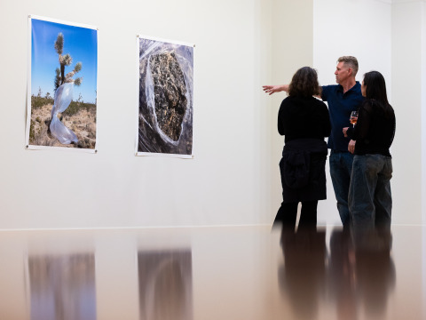 A man wearing a navy polo shirt is gesturing towards two large photographs on a white wall as he explains them to two women wearing black shirts.
