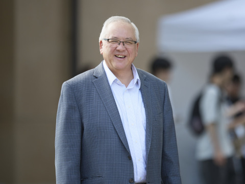 SUA President Ed Feasel is wearing a grey suit jacket, white shirt, glasses as he smiles at the camera