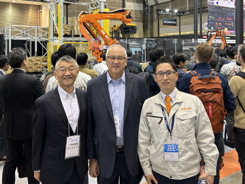 Two men wearing suit jackets and dress shirts and a third man wearing a jacket with "Mujin" embroidered on it smile and pose for a photo together at the 2025 International Robot Exhibition in Tokyo.