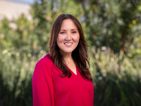 A professional headshot of a smiling woman with long brown hair, wearing a vibrant magenta V-neck blouse. She is positioned against a soft-focus background of lush green foliage and trees.
