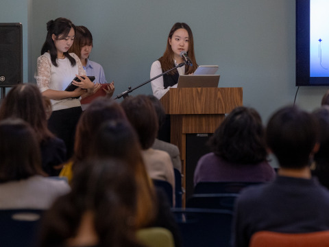 Three people wearing professional attire stand at the front of the room. One is speaking into a microphone at a wooden lectern to a seated audience.