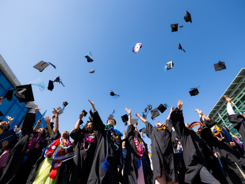 SUA Graduates wearing a mix of colorful clothes under their black graduation gowns throw their caps into the air in celebration.