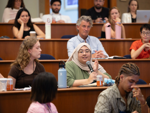 A woman wearing a tan headscarf and green shirt speaks into a microphone as people sitting around her listen on.