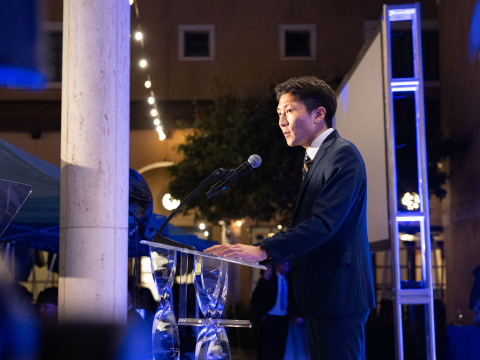 Side view of a man with short black hair wearing a black suit speaking into a microphone at a clear lectern with glass legs during the 2026 Peace Gala.