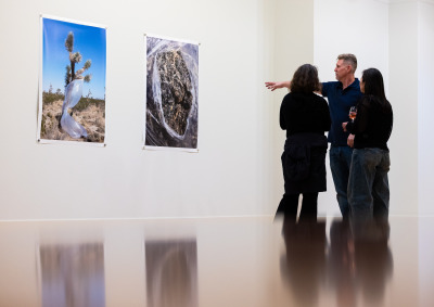 A man wearing a navy polo shirt is gesturing towards two large photographs on a white wall as he explains them to two women wearing black shirts.
