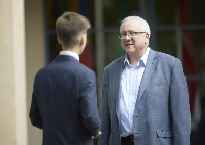 SUA President Ed Feasel is wearing a grey suit jacket and white shirt. He smiles as he speaks to a student wearing a blue suit.