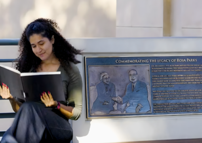 A woman with curly brown hair looks at a large black book as she sits beside a plaque commemorating Rosa Parks' legacy.