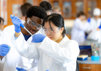 A male and female student in lab coats and safety goggles work together with a micropipette in a science lab.
