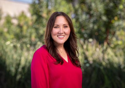 A professional headshot of a smiling woman with long brown hair, wearing a vibrant magenta V-neck blouse. She is positioned against a soft-focus background of lush green foliage and trees.
