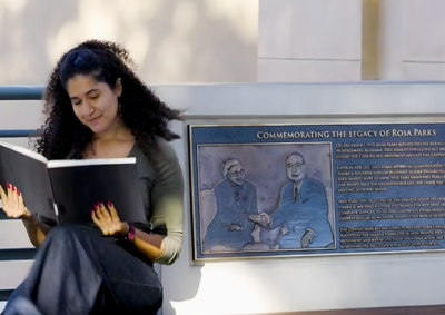 A woman with curly brown hair looks at a large black book as she sits beside a plaque commemorating Rosa Parks' legacy.