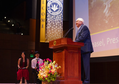 President Feasel wears a suit and speaks into the microphone from behind a wooden lectern. Two students are seated off to the left.