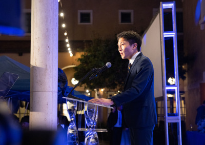 Side view of a man with short black hair wearing a black suit speaking into a microphone at a clear lectern with glass legs during the 2026 Peace Gala.