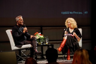 Simon Mainwaring and Gail Becker speak into microphones while sitting in white chairs on stage in the Soka Black Box Theatre